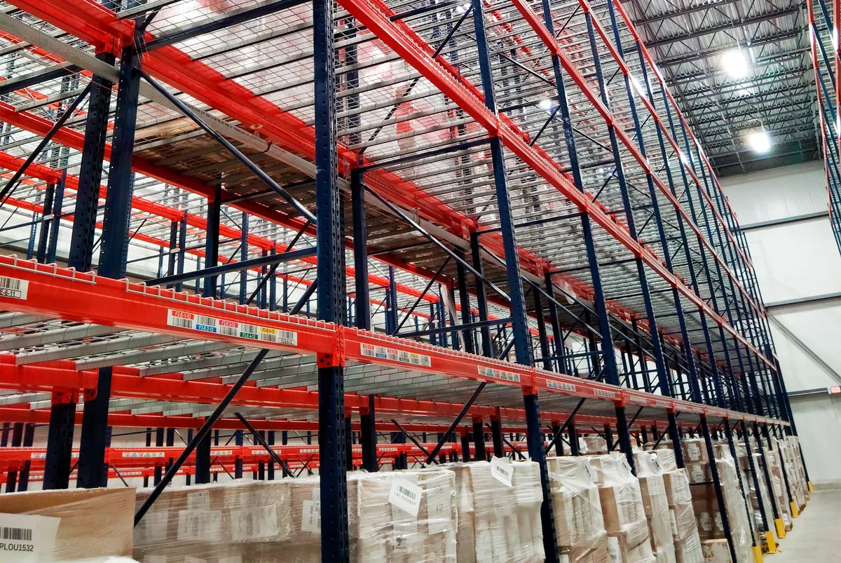 Tall pallet racking in the storage hall, each bay barcode-labeled by location, with shrink-wrapped pallets of cartons staged at floor level